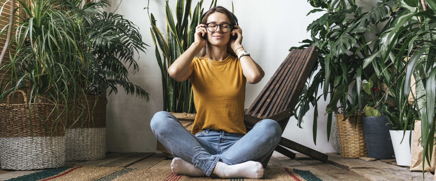 Frau sitzt zwischen Zimmerpflanzen auf einem Teppich und h&ouml;rt Musik